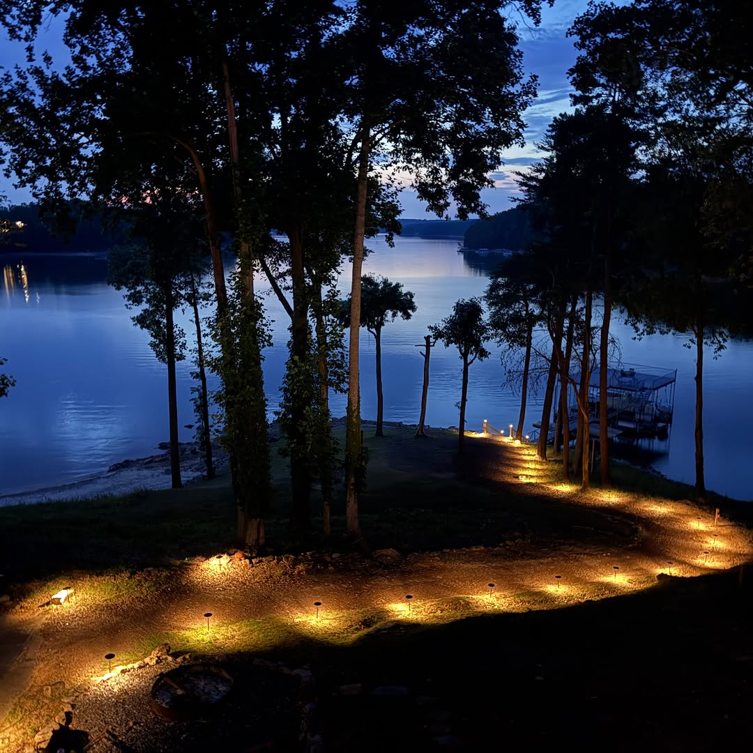 Outdoor path lights leading through trees beside a lake at dusk